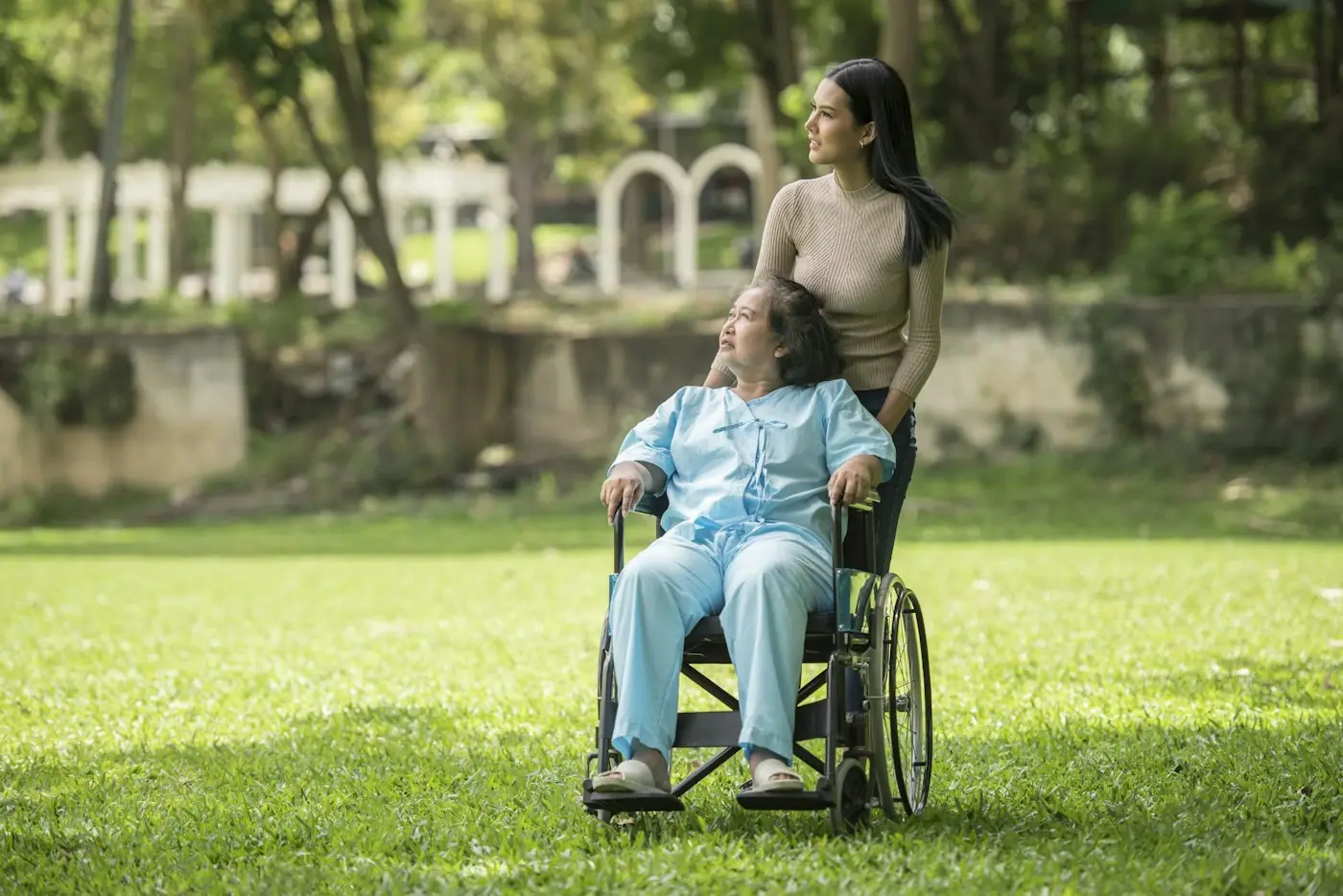 Older man in a wheelchair enjoying a park with a younger woman standing behind him
